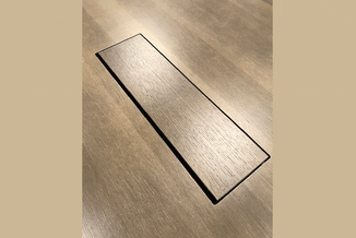 Grijze PVC-plank met houtnerf-oppervlak op laminaatvloer met houtstructuur, gezien vanuit schuin perspectief.