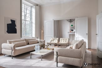 Moderne woonkamer met beige stoffen banken, witte deuren, glazen salontafel, ronde fauteuils en abstracte wandkunst.