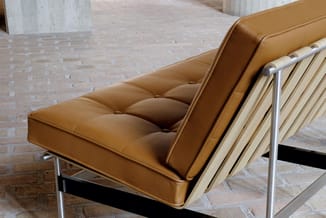 Bruine leren design zetel met gestoffeerde zitting, metalen frame en armleuningen, in kantoorruimte met betonnen pilaren.