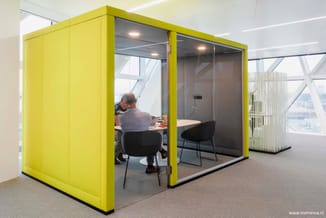 Gele geluidwerende belcel met glazen wanden in kantoor, voorzien van tafel, zwarte stoel en verlichting.