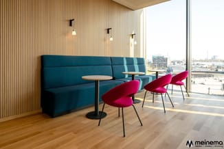 Moderne kantoorruimte met blauwe modulaire zitbank, roze ergonomische stoelen, ronde tafels en houten lambrisering tegen pano
