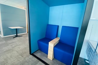 Blauwe belcel met twee zitplaatsen, houten tafeltje in het midden, lichtblauwe achterwand, kantooromgeving.