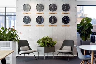 Kantoorruimte met acht wereldklokken aan betonnen wand, twee zwarte stoelen met grijze zitting, plantenbakken, ronde tafels e