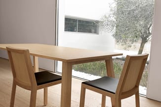 Houten eettafel met twee houten stoelen met zwarte zitting in moderne kantoorruimte met grote ramen en uitzicht op tuin.