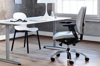 Modern kantoor met wit bureau, witte designstoel met zwarte poten, grijze ergonomische bureaustoel op wieltjes, betonlook wan