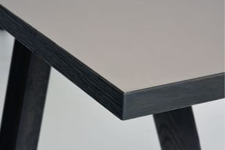 Hoekdetail van tafel met grijs blad en zwart gelakt houten frame tegen witte wand.