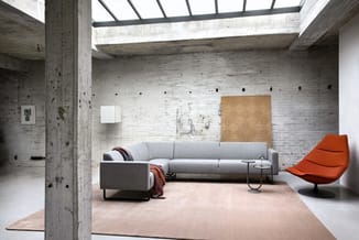 Moderne lounge met grijze hoekbank, oranje fauteuil, betonnen kolommen, witte bakstenen muur en natuurlijk daglicht van boven