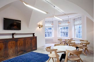 Modern kantoor met houten ronde tafels, lichte houten stoelen, houten dressoir, TV-scherm, blauwe tapijt en grote ramen met r