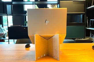 HP-laptop in geel/gouden laptopstandaard van meinema.nl op houten bureau in moderne kantoorruimte met zwarte kastenwand.