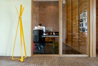 Gele kapstok in minimalistisch design met drie poten, geplaatst in moderne kantoorruimte met glazen wand en bakstenen muur.