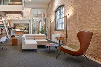 Moderne kantoorlounge met cognackleurige lederen fauteuil, beige modulaire bank, kleine tafel en houten accent, bakstenen wan