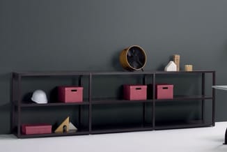 Donkergrijs open kastensysteem met roze opbergboxen, decoratieve objecten en een ronde wandlamp tegen grijze muur.