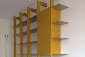 Gele open boekenkast met donkergrijs metalen planken tegen witte muur, met staande lamp links.