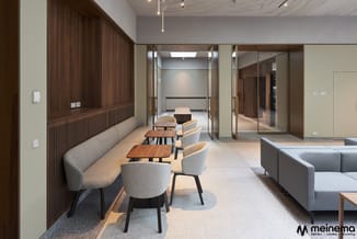 Moderne kantoorlounge met grijze fauteuils, houten tafels, lange grijze zitbank tegen donkerhouten wandpaneel en grijze loung