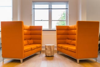 Twee oranje loungezetels met hoge ruggen en houten poten tegenover elkaar, tussen een houten bijzettafel, voor grote ramen in