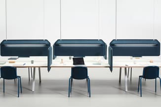 Drie werkplekken met witte bureaus, donkerblauwe stoelen en hangend akoestische schermen boven elke werkplek.