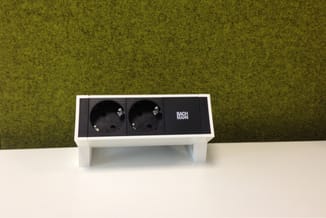 Witte Bureau-stekkerdoos BACHMANN met twee zwarte stopcontacten en één zwart vak, op witte werkblad tegen groen-geel gemêleer