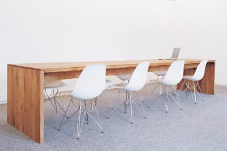 Houten vergadertafel met witte kunststof stoelen op metalen poten in kantoorruimte met grijze vloer.