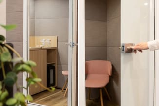 Kantoor belcel met glazen deur, houten werkblad met accessoires, roze stoel en grijze wandtegels.