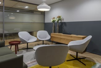 Moderne kantoorlounge met grijze fauteuils, houten dressoir, rode bijzettafel, gele vloermat en glaswanden.