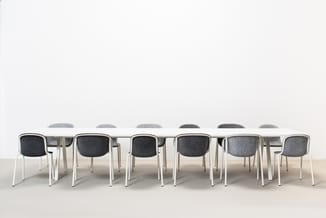 Vergadertafel met acht grijze stoelen met witte metalen poten in minimalistische ruimte met witte muren.