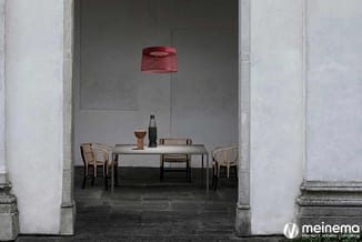 Minimalistisch ingerichte ruimte met grijze tafel, houten stoelen, rode hanglamp en decoratieve objecten tegen betonnen wand.