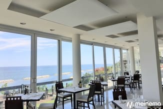 Modern restaurant met zwarte stoelen aan witte tafels, grote raampartijen met zeezicht, betonnen kolommen en plafond met inbo