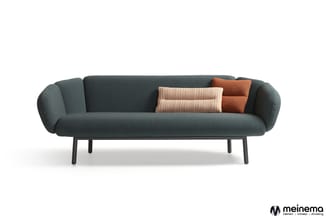 Grijsgroene bank met afgeronde vormen, zwart metalen poten, twee decoratieve kussens in beige en terracotta.