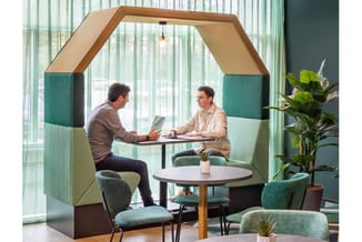 Akoestische werkplek met houten frame en groene/beige bekleding. Twee mannen zitten achter tafel in modern kantoor met raamvi