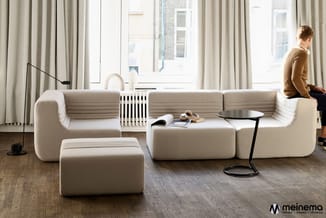 Modulaire beige-grijze zithoek in moderne ruimte met grote ramen, houten vloer, staande lamp en zwarte bijzettafel. Meinema-l