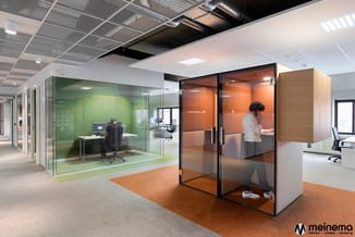 Moderne kantoorruimte met glazen belcellen in groen en oranje, werktafels, bureaustoelen en onderverdeelde werkplekken onder