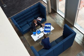 Twee personen in gesprek op blauwe modulaire zitgroep rond witte tafel met documenten in modern kantoorinterieur.