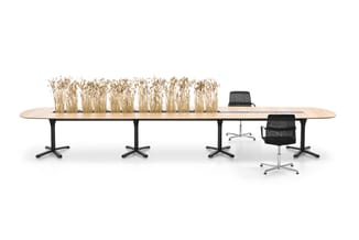 Lange houten vergadertafel met zwarte stalen poten, bezet met droogbloemen. Zwarte bureaustoel ernaast. Minimalistische kanto
