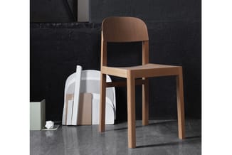 Houten stoel met natuurlijke afwerking en gebogen rugleuning tegen zwarte muur, naast stapel witte doeken op grijze vloer.