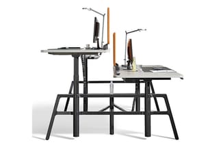 Ergonomisch werkstation met twee zwarte bureaubladverhogers op metalen frame, houten armleuningen en verstelbare monitorarmen