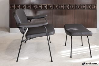 Zwarte leren fauteuil met armleuningen en metalen frame naast bijpassende zwarte voetenbankje in minimalistisch interieur.