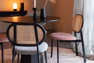 Ronde zwarte eettafel met twee stoelen: een met rotan rug en grijze zitting, een met rotan rug en roze zitting. Warm verlicht