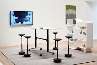 Modern kantoorinterieur met staande werkplek, zwarte zit-sta krukken, witte kastenwand, TV-scherm aan muur en houten vloer.