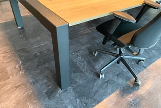 Houten bureau met grijze metalen onderstel en zwarte bureaustoel met wielen op donker tegelvloer in kantoorruimte.