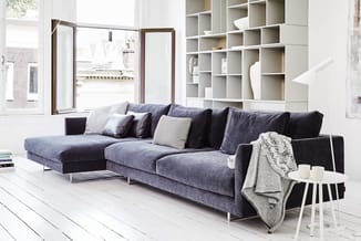 Grijsblauwe hoekbank met chaise longue, zwarte zijkussen en beige plaid in lichte, moderne kamer met witte vloer en inbouwkas