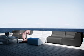 Modulaire loungebanken in grijs en wit met blauwe poef op houten terras aan zee, naast zwarte bijzettafel met glazen.