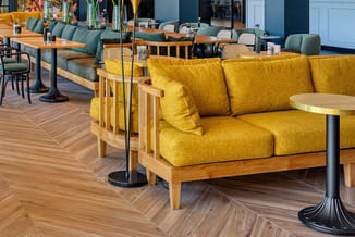 Lounge-ruimte met gele fauteuils met houten frame, groene banken op de achtergrond, houten visgraat vloer en ronde bijzettafe