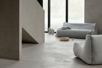 Minimalistisch interieur met grijze zitzakken, grijze rollen stof, houten schaal en betonnen elementen bij grote ramen.