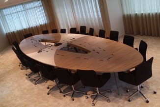 Grote houten vergadertafel met organische vorm, zwarte bureaukstoelen, grijze verticale lamellen achter, betonnen vloer.