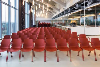 Rood vergaderzaal met rijen rode kunststoftoelen met metalen poten, grote ramen, hangende verlichting en glazen kantoorruimte