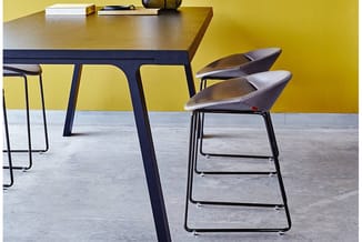 Moderne werkplek met houten tafel op donkerblauw onderstel, grijze barkrukken met zwart frame, gele en grijze muurafwerking.