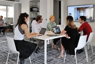 Vijf personen zitten rond een witte vergadertafel met laptops in een moderne kantoorruimte met witte stoelen en grijs tapijt.