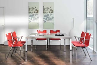 Vergaderruimte met grijze tafel, zes rode stoelen met armleuningen, witte wanden, houten vloer en twee groene wandpanelen.
