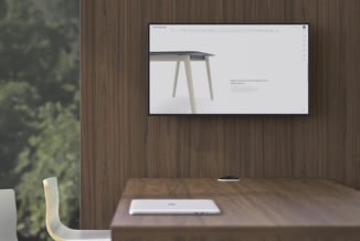 Houten kantoor met wandmontitor waarop designtafel met donkergrijs blad en lichte poten wordt getoond. Bureaulade op houten t