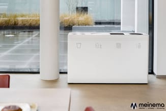 Witte afvalbak met vier gescheiden compartimenten en recyclingpictogrammen op een kantoorbureau bij een raam met uitzicht op